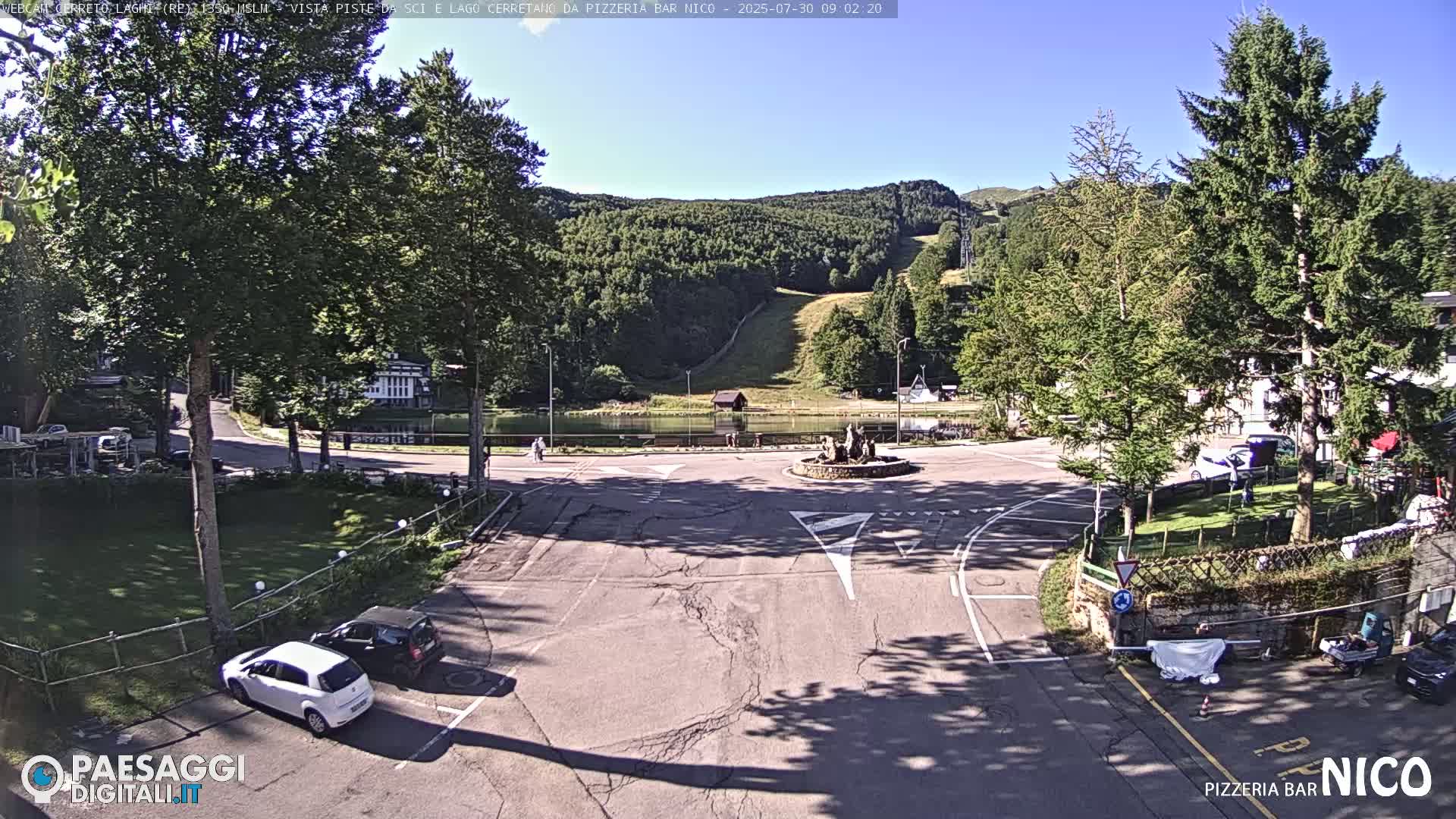 A sunny day reveals a mountainside with a ski slope, a small lake, and a parking area with several cars near a circular intersection, all surrounded by lush green trees.