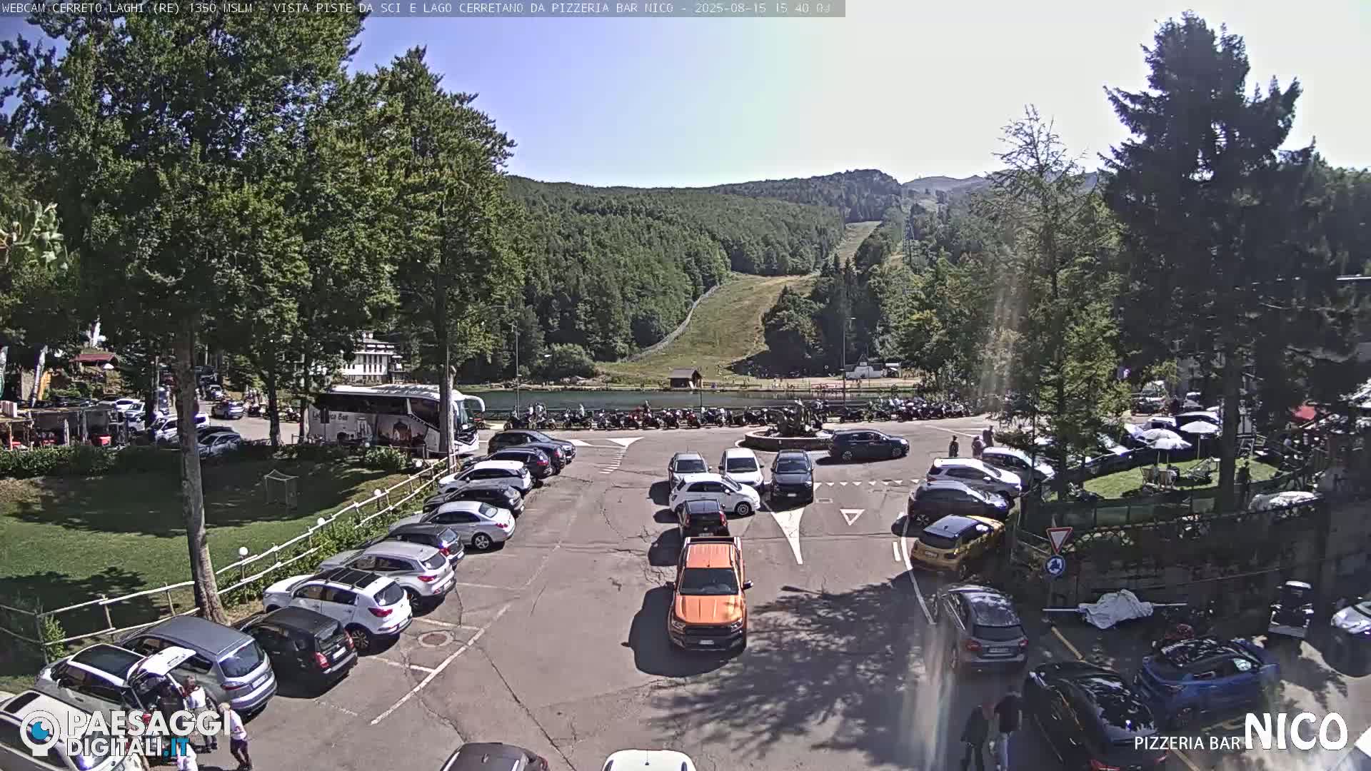 A sunny day reveals a mountainside with a ski slope, a small lake, and a parking area with several cars near a circular intersection, all surrounded by lush green trees.