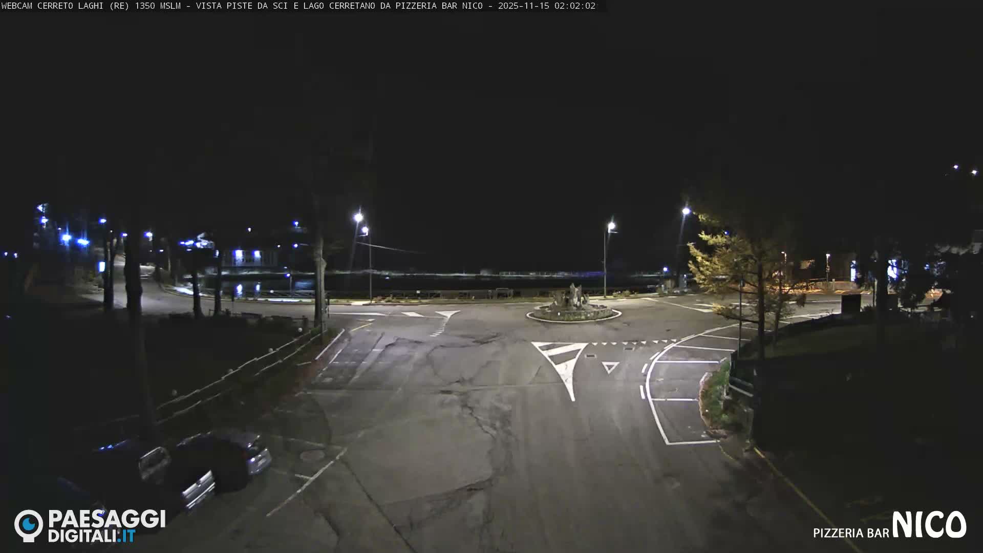 A clear night view reveals a quiet road intersection with white markings and a central island sculpture, illuminated by streetlights, with a dark body of water and distant lights visible in the background under a clear sky.
