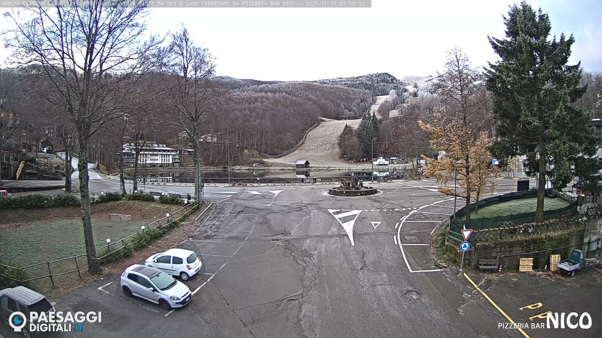A clear night view reveals a quiet road intersection with white markings and a central island sculpture, illuminated by streetlights, with a dark body of water and distant lights visible in the background under a clear sky.