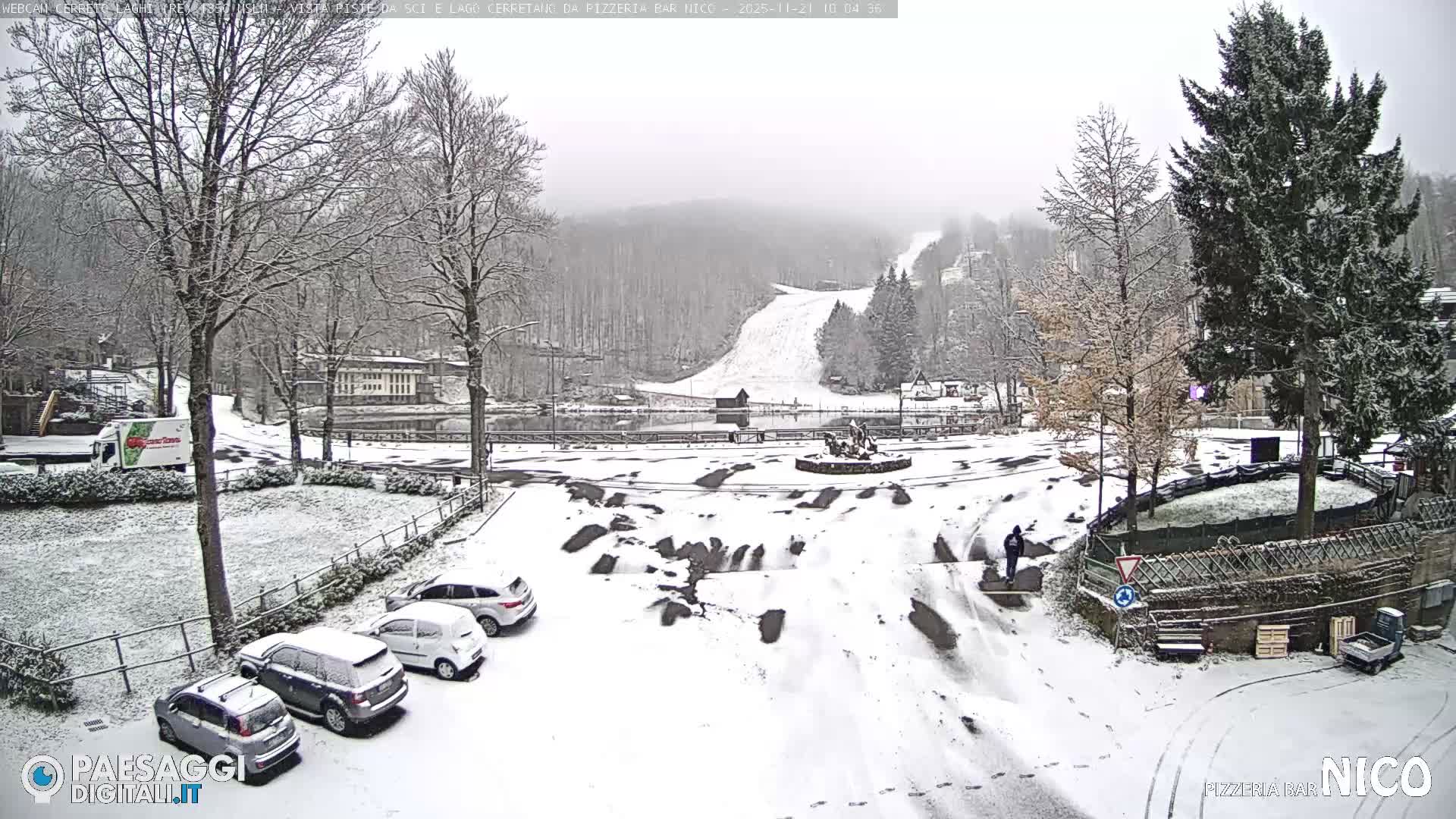 A snow-covered mountain village is captured under an overcast sky, featuring freshly powdered trees, roads, and cars, a central lake, and a distant ski slope, with a person walking through the wintry scene.