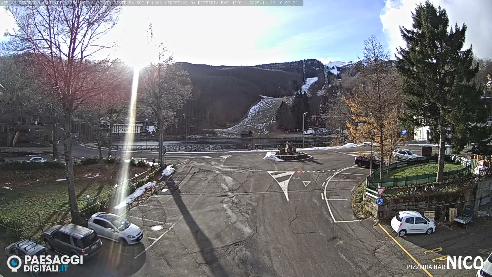A bright, sunny winter day with scattered clouds illuminates a cracked asphalt roundabout with a small fountain and several parked cars in the foreground, overlooking a tranquil lake and a densely forested mountain with a partially snow-covered ski slope, leading up to distant snowy peaks.