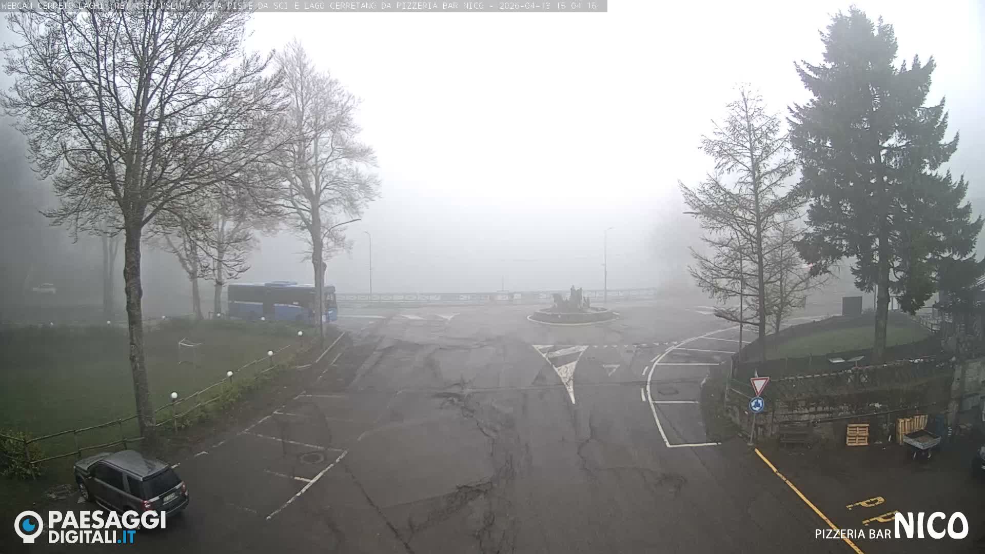 A foggy day obscures a mountain road and surrounding trees, with several cars parked on the dark asphalt, which has patches of old snow near the roadside.