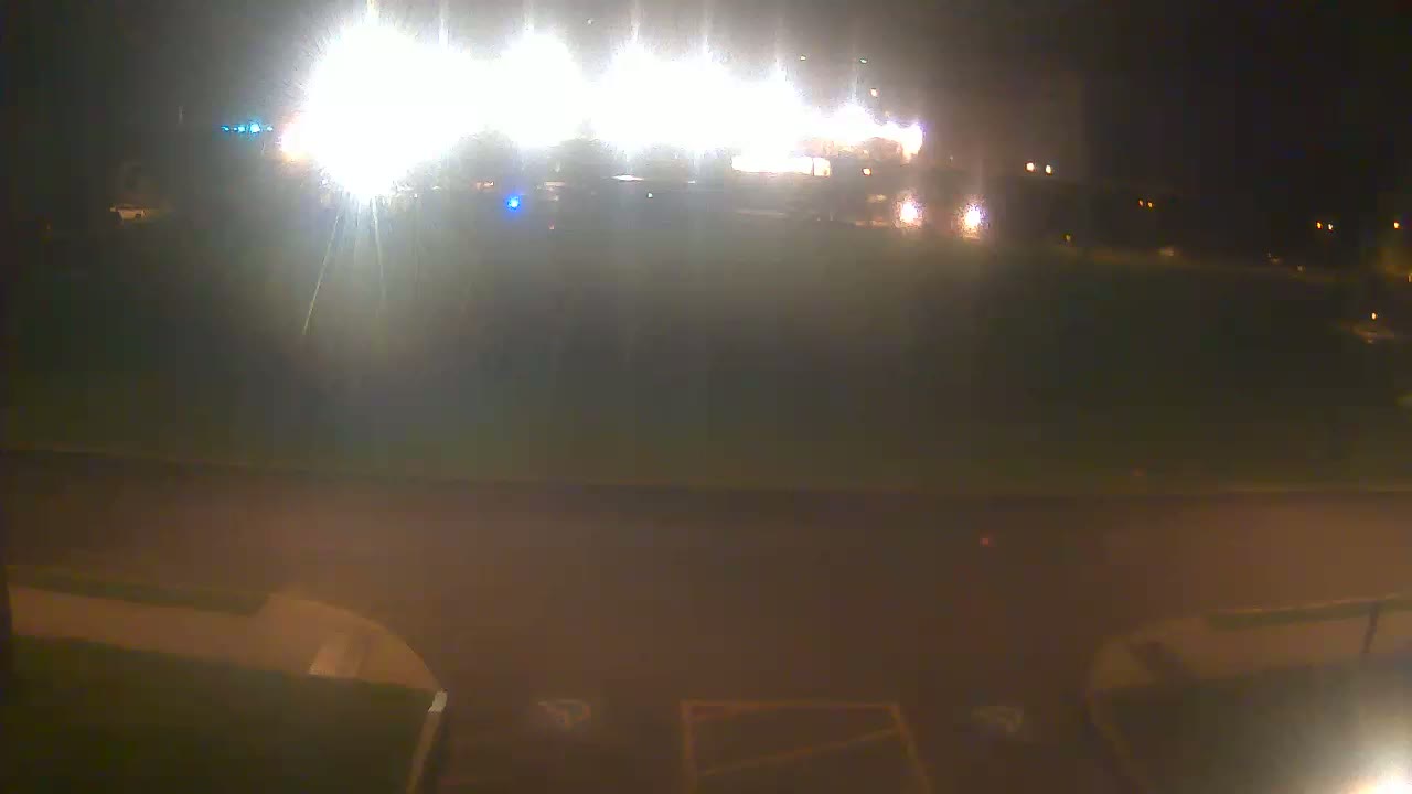 A brightly lit building is visible at night across a dark, empty field, from a low vantage point near the ground.