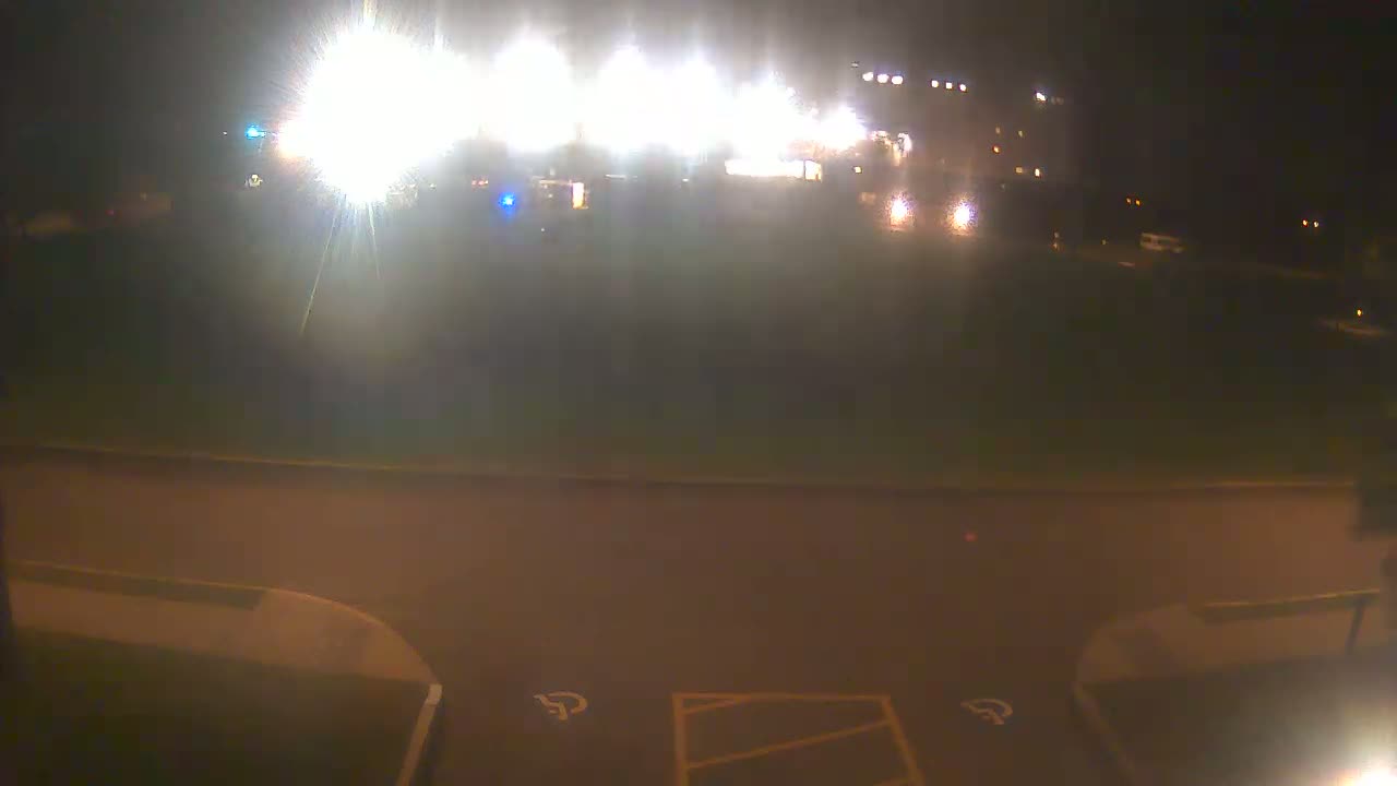 At night, brightly lit vehicles and a building are visible in the distance, with a dark, empty parking lot in the foreground.
