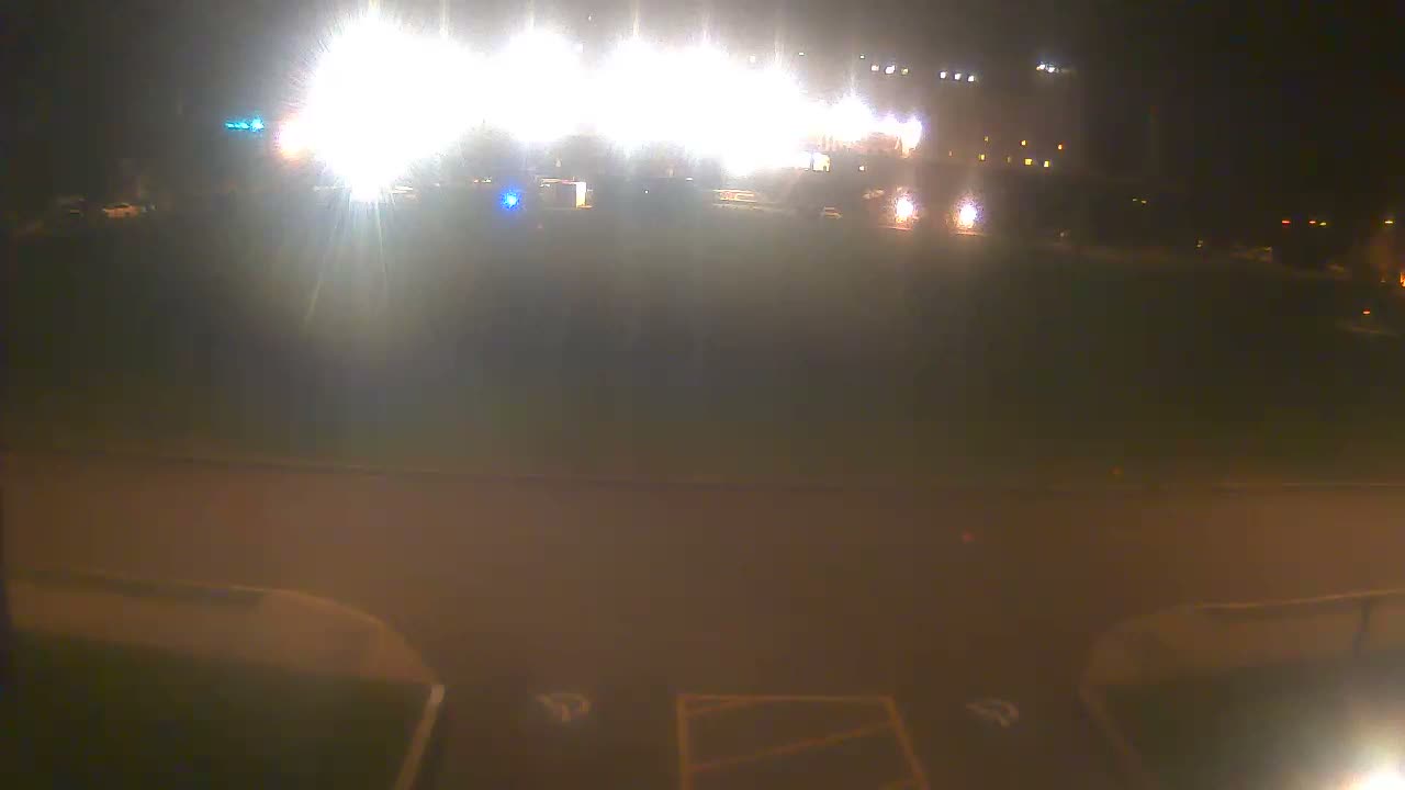 A nighttime outdoor scene displays intense glare from numerous bright artificial lights illuminating a dark field or open ground, with a paved area featuring faint markings in the foreground, under clear conditions.