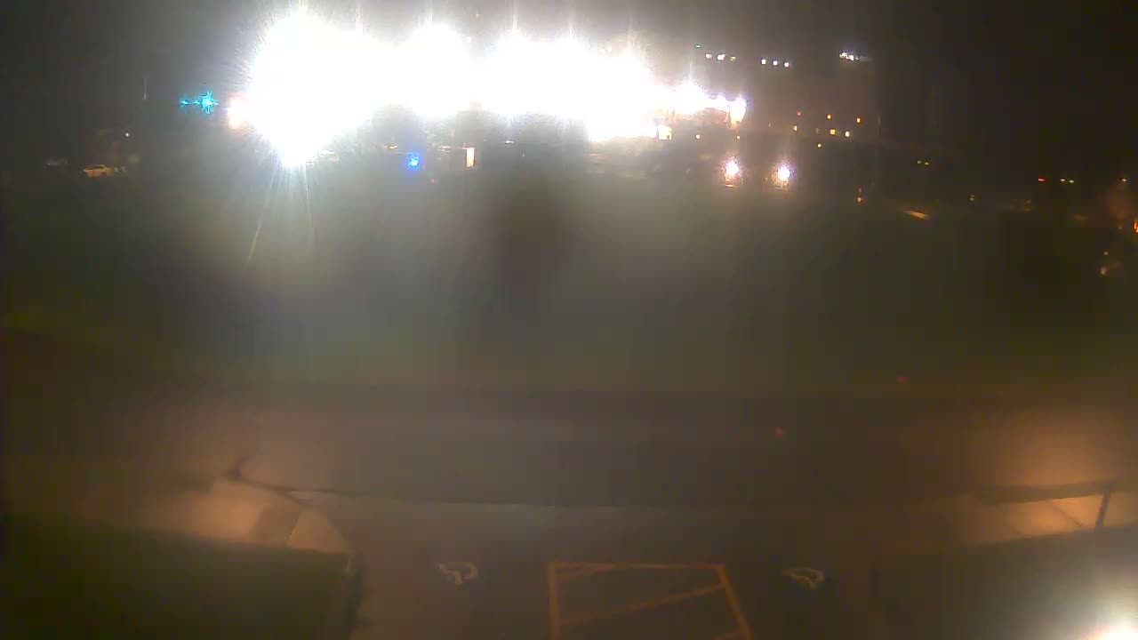 A very bright and hazy outdoor night scene reveals intense artificial white, blue, and green lights reflecting vividly on a wet ground, indicating rain or damp conditions.