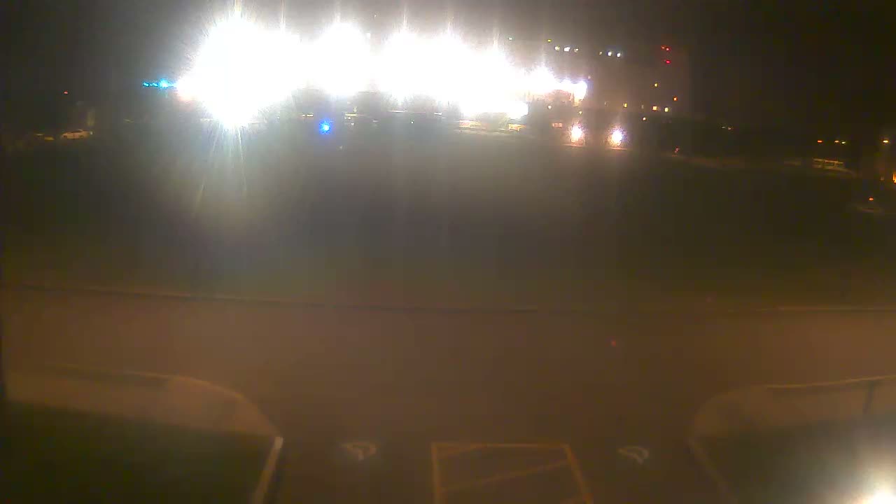 A dark outdoor scene at night is illuminated by numerous intense, diffused bright lights, some appearing to be from emergency vehicles, casting a significant glare over a paved foreground and distant blurred structures.
