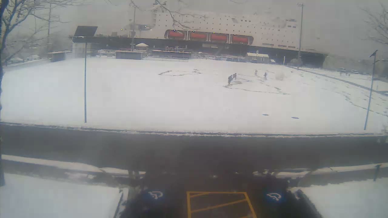 A dark outdoor scene at night is illuminated by numerous intense, diffused bright lights, some appearing to be from emergency vehicles, casting a significant glare over a paved foreground and distant blurred structures.