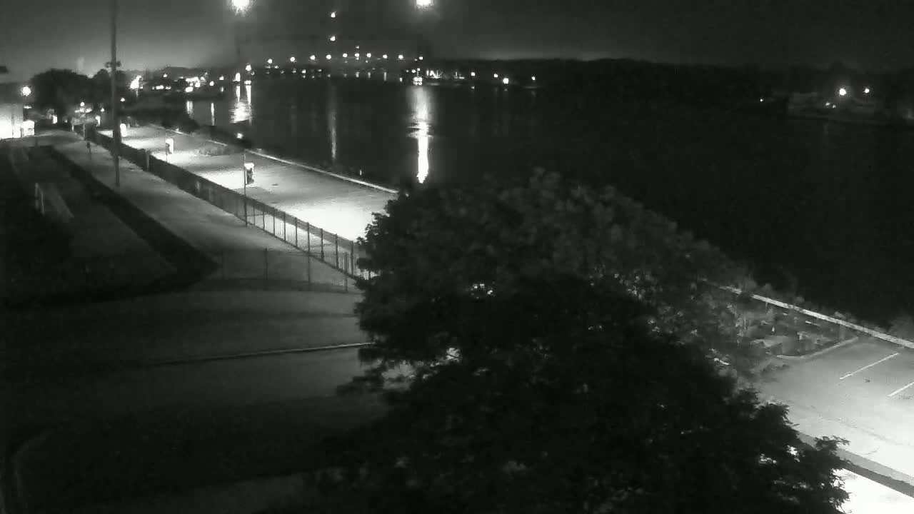 A nighttime, high-angle view shows a paved walkway along a calm body of water, with a tree and some lights visible.