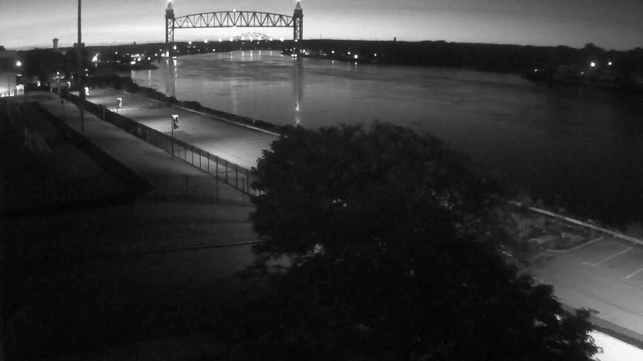 A dark, nighttime view shows a wide river with a large metal bridge in the distance,  a paved path along the riverbank, and trees in the foreground.