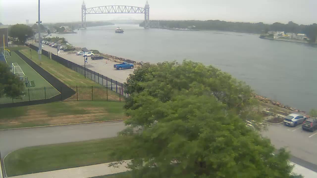 A wide shot shows a body of water with a tall bridge in the distance on an overcast day, with a small athletic field, parking lots, and trees in the foreground.