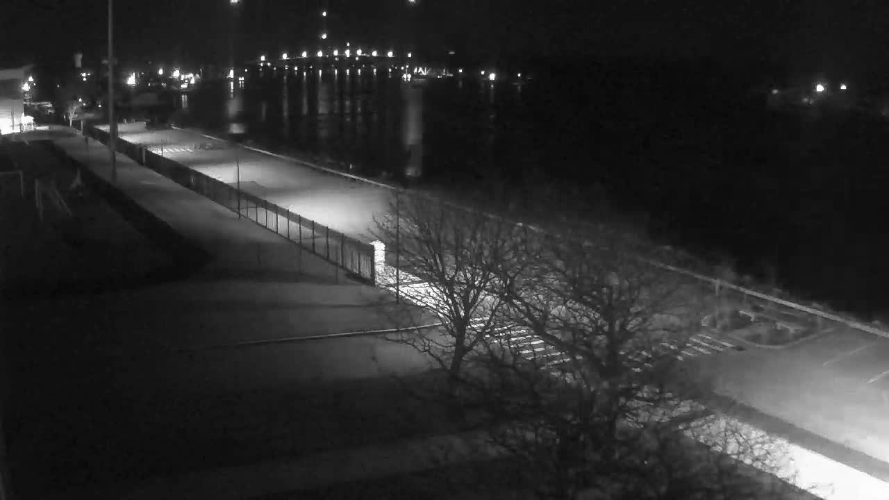 A nighttime view overlooks a calm body of water reflecting distant city lights, with a paved waterfront path, bare trees, and streetlights in the foreground under clear skies.