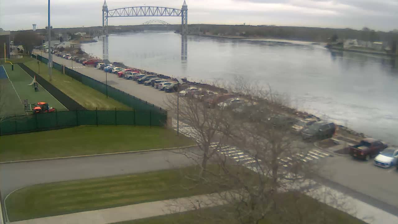 On an overcast day, a wide river or canal with a distant blue truss bridge is visible, bordered by a parking lot filled with cars, a paved road, and a sports complex featuring a green field and track.