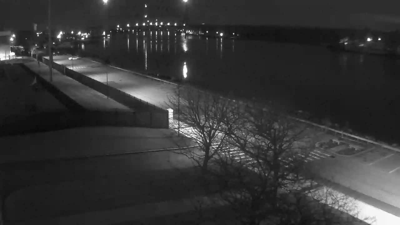 On a clear night, a black and white, high-angle view captures a calm river reflecting numerous distant lights, running parallel to a street with a single parked car and bare trees in the foreground.