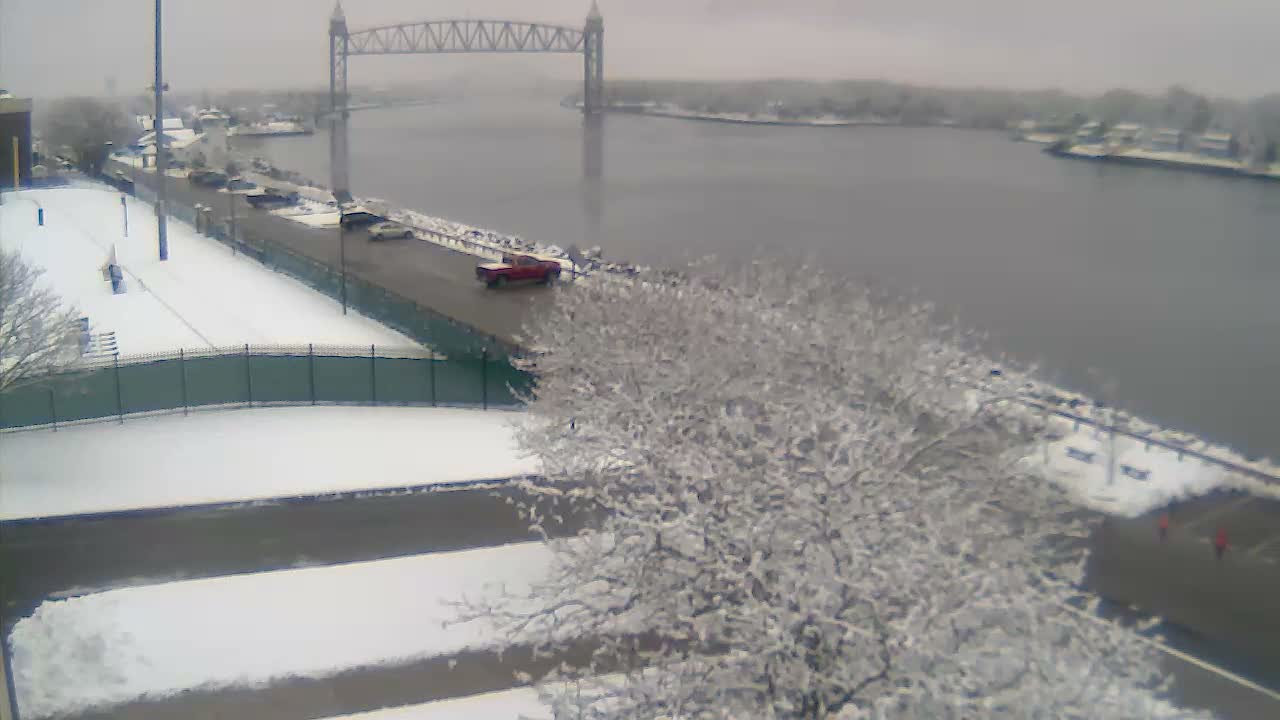 On a clear night, a black and white, high-angle view captures a calm river reflecting numerous distant lights, running parallel to a street with a single parked car and bare trees in the foreground.