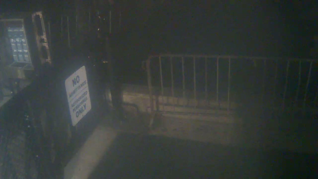 A dark, possibly nighttime scene shows a metal barricade next to a wall with a keypad and a sign.