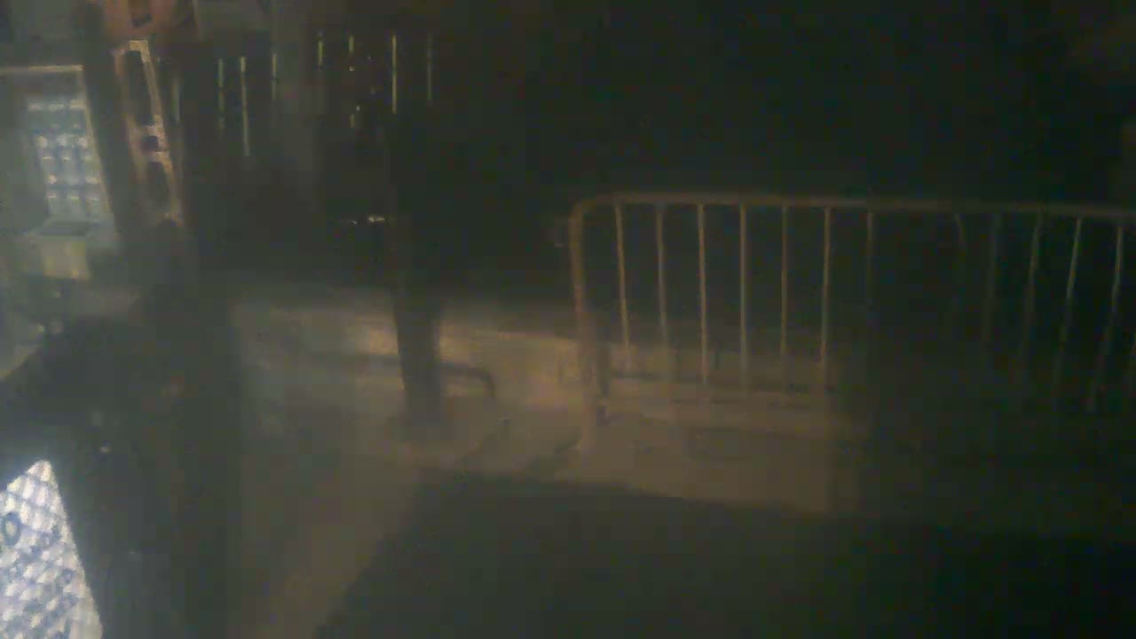 A dark, blurry outdoor night scene shows a light-colored metal barrier on the right, subtle vertical lights on the left, and a reflective ground suggesting damp conditions.