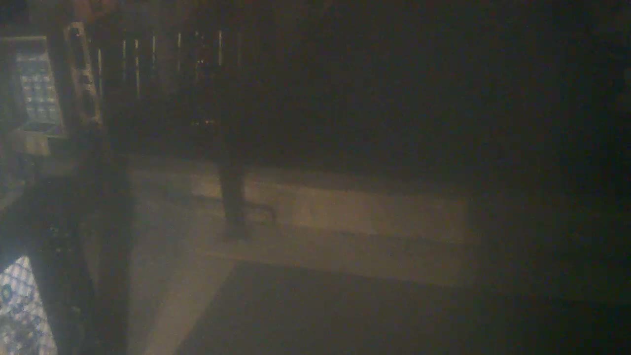 A blurry and very dark night image shows a person standing on a paved surface near a low wall, with urban structures faintly visible in the background.