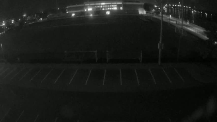 A nighttime, low-light view shows an empty sports field with a parking lot in the foreground and a building in the background.
