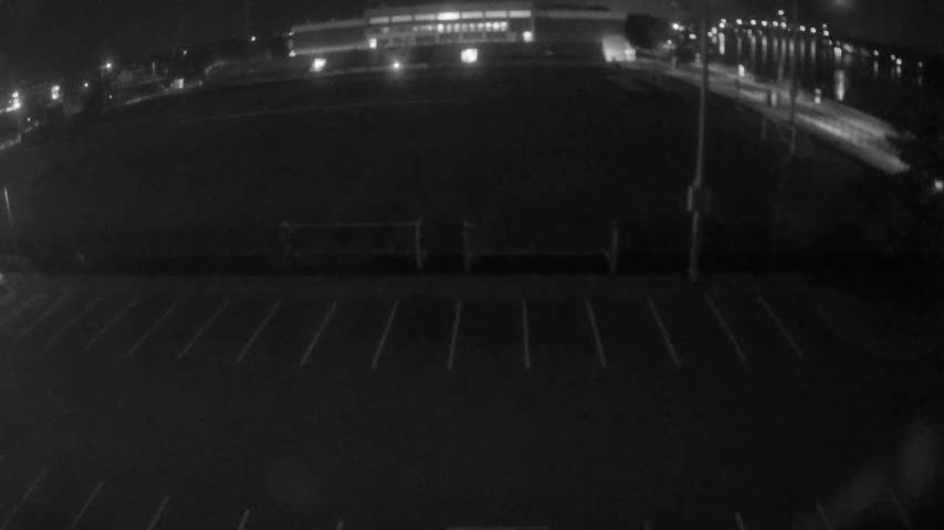 A nighttime, low-angle view shows an empty parking lot in front of a large, dark building, next to a body of water with lights visible across it.