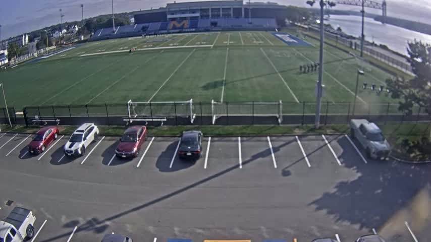Several cars are parked in a lot next to a football field and bleachers, with a body of water and a bridge visible in the distance on an overcast day.