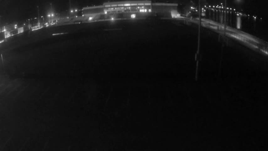 The image depicts a dimly lit outdoor scene at night, featuring a wide dark expanse in the foreground, a brightly lit building and streetlights in the background, and a body of water with glowing reflections and a lit structure to the right, all under clear skies.