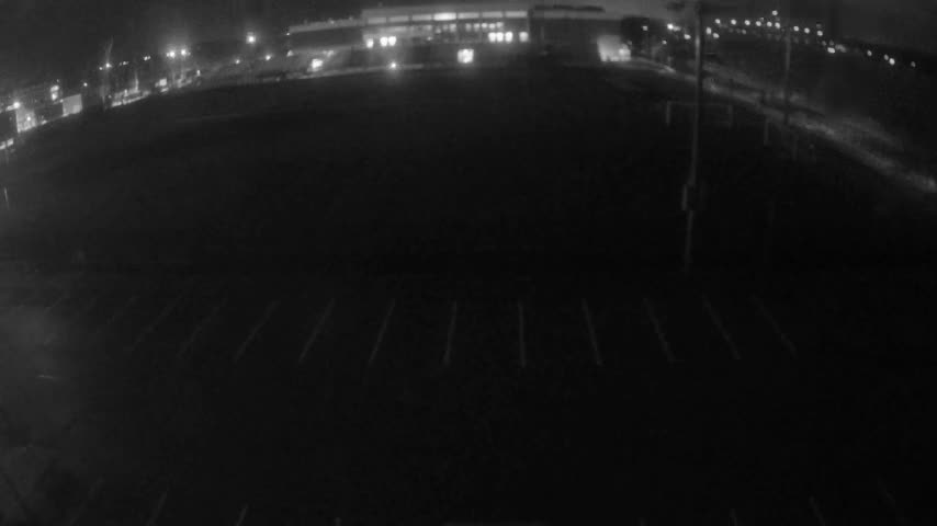 An extremely dark, wide-angle outdoor nighttime image shows an empty paved area with faint diagonal parking lines in the foreground, a brightly lit building in the distant background, and a curving road lined with numerous streetlights on the right, under clear night skies.