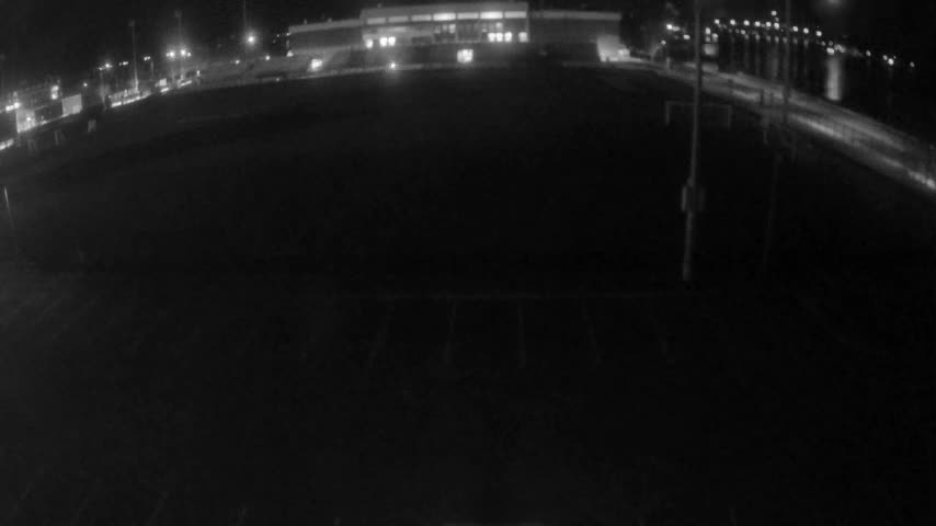 An extremely dark, wide-angle outdoor nighttime image shows an empty paved area with faint diagonal parking lines in the foreground, a brightly lit building in the distant background, and a curving road lined with numerous streetlights on the right, under clear night skies.