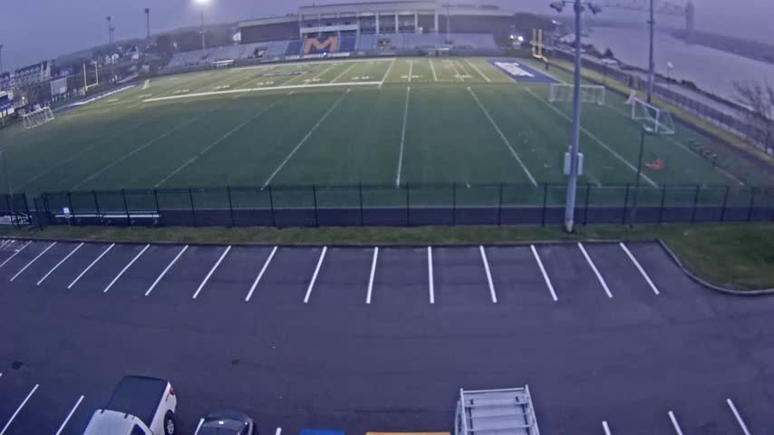 A dark nighttime, wide-angle view captures a wide, unlit field in the foreground, with an illuminated building in the distance and a lighted bridge extending over water to the right.