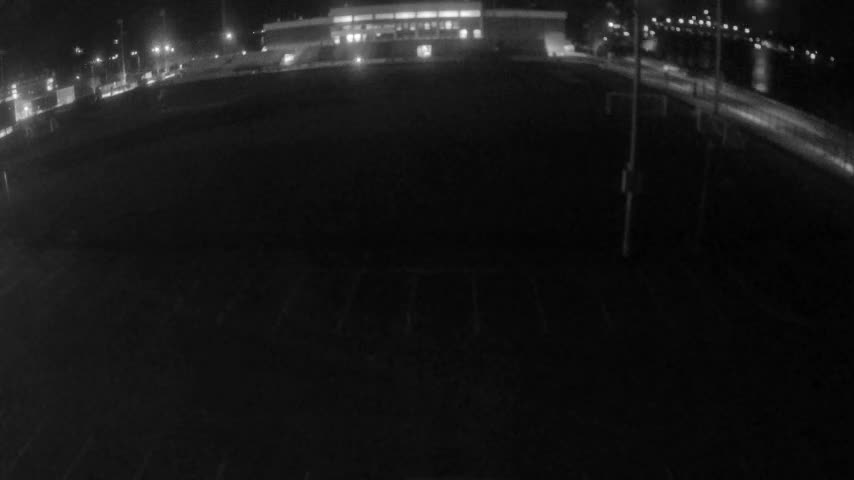 A wide-angle, low-light night view captures an empty outdoor field with faint markings in the foreground, set against a backdrop of a brightly lit building and a waterfront road, all under dark, rainy skies.