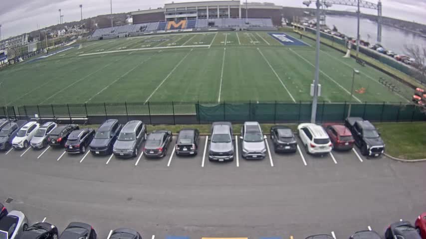 Under an overcast sky, an outdoor scene shows a parking lot full of cars in the foreground, a green stadium football field and bleachers in the middle, and a river with a bridge in the background.