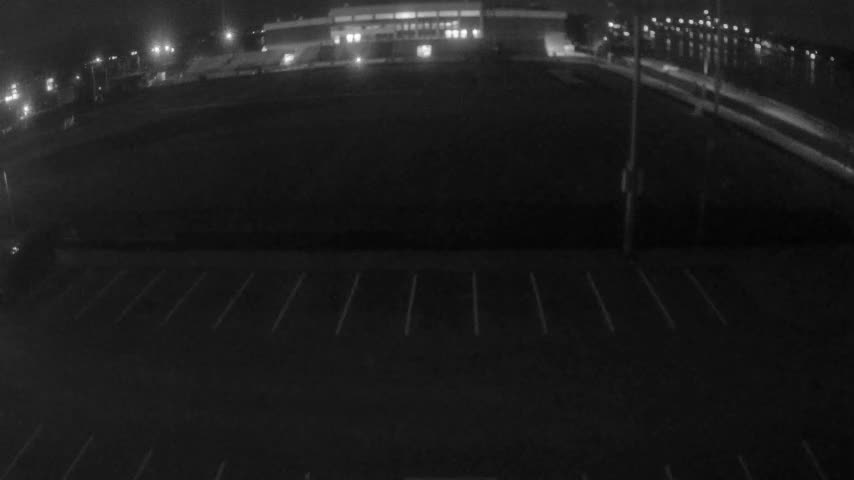 An aerial night view captures an empty parking lot in the foreground, leading to a dark field or open area, with a brightly lit building and a line of distant lights visible under a clear night sky.
