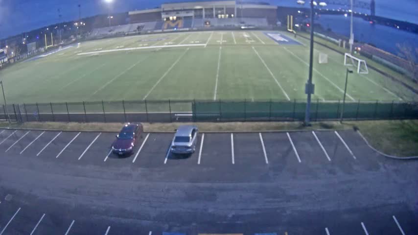 An aerial night view captures an empty parking lot in the foreground, leading to a dark field or open area, with a brightly lit building and a line of distant lights visible under a clear night sky.
