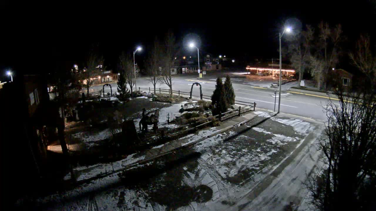 The image displays a quiet town street at night illuminated by streetlights and distant building lights, with a landscaped median containing bare trees and a fence, all under clear dark skies.