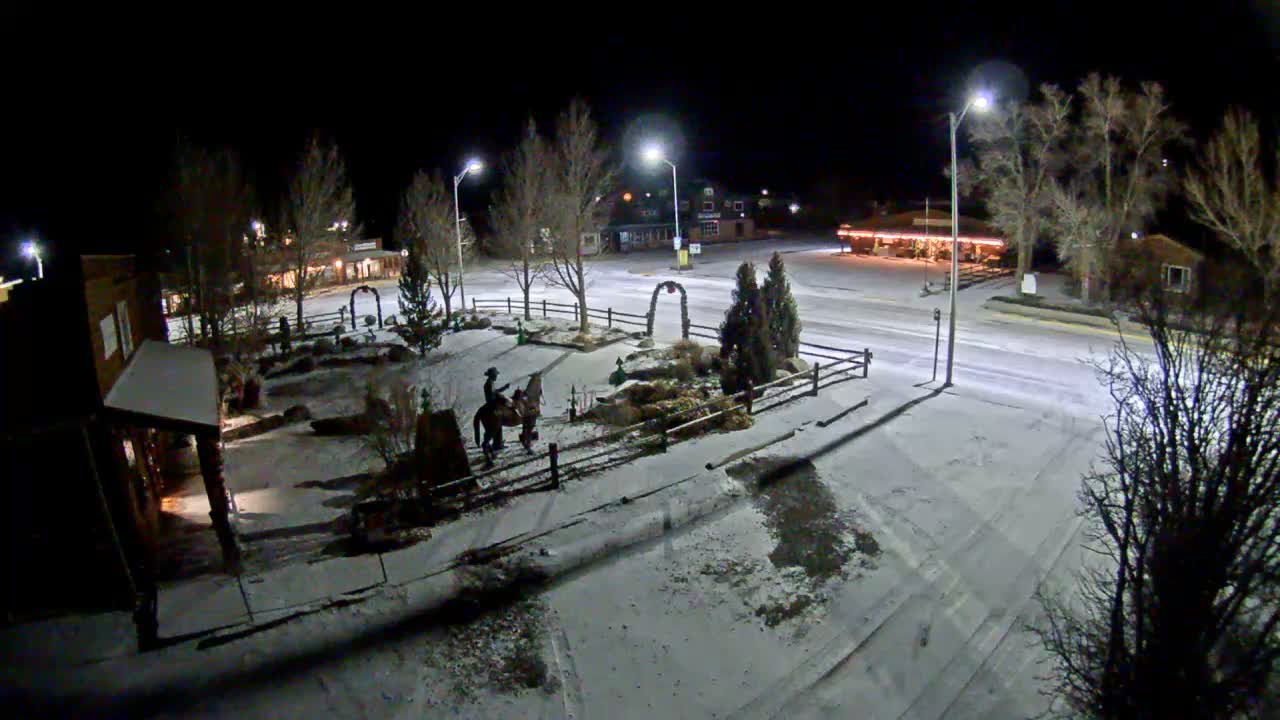 A clear, cold winter night illuminates a snow-dusted small town square, revealing bare trees, streetlights, buildings with warm interior glows, and a prominent statue of a person on horseback.