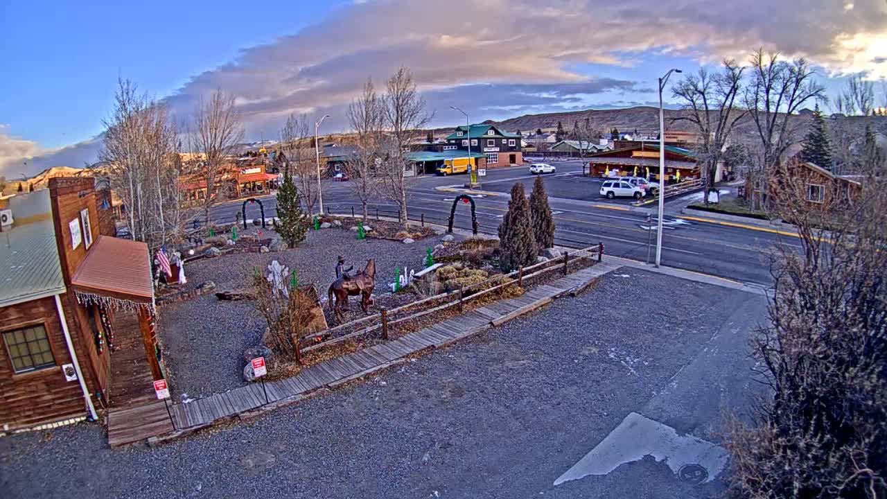 An elevated view reveals a small town scene with a gravel foreground featuring a person on a donkey statue and sparse winter trees, across a street lined with commercial buildings, all set against distant hills under a partly cloudy sky.
