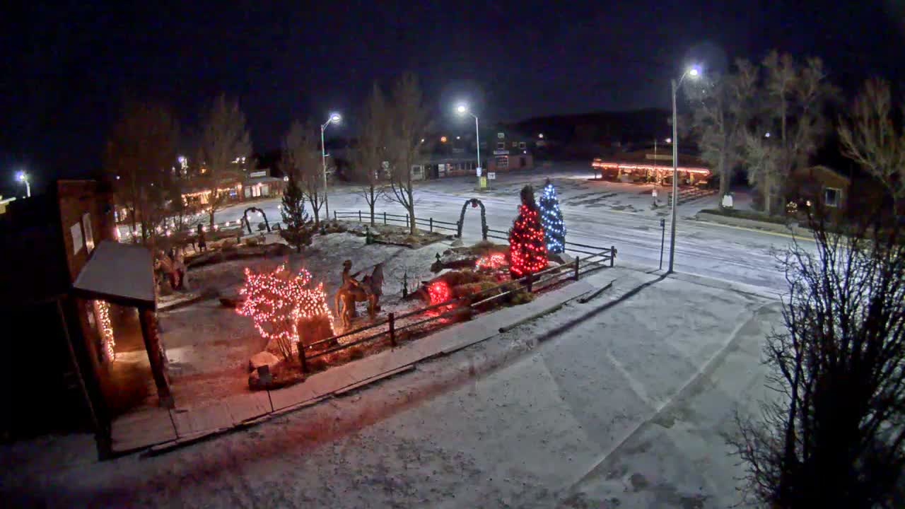 A snowy town square is illuminated at night by streetlights and festive red, blue, and white Christmas lights adorning trees and a cowboy statue, under clear, cold winter conditions.