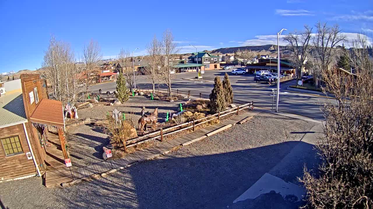 A snowy town square is illuminated at night by streetlights and festive red, blue, and white Christmas lights adorning trees and a cowboy statue, under clear, cold winter conditions.