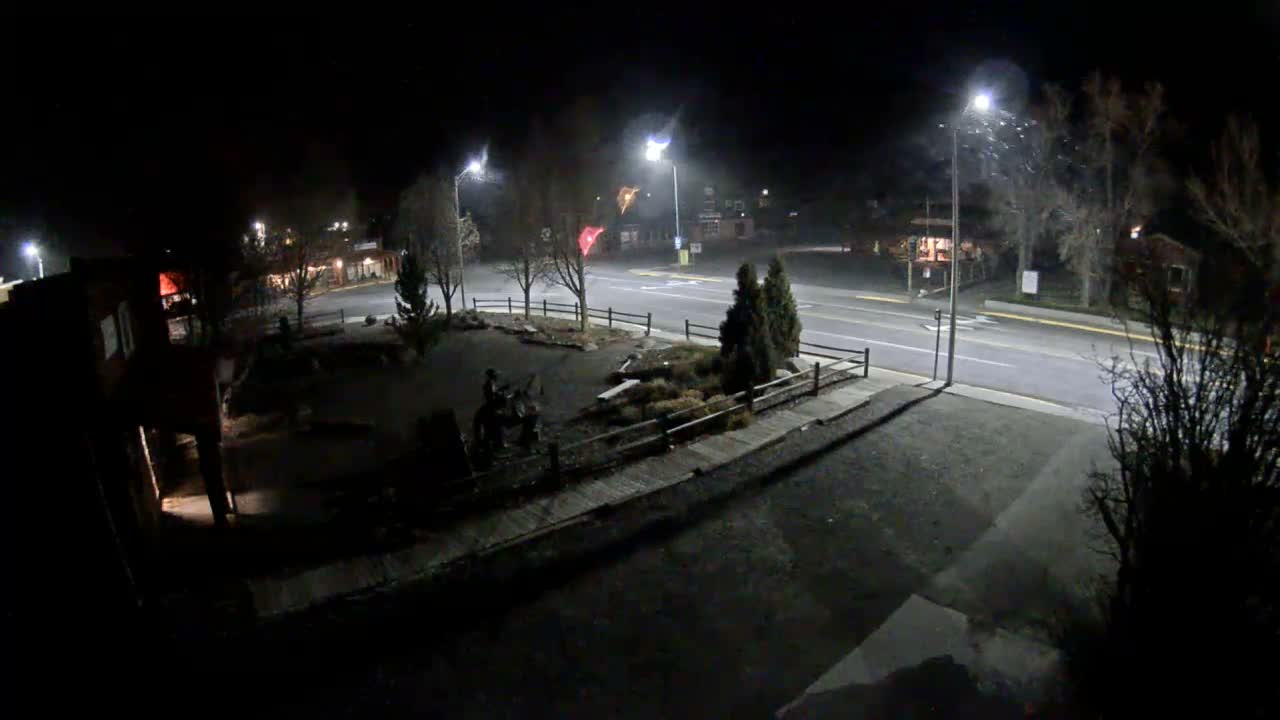 A snowy town square is illuminated at night by streetlights and festive red, blue, and white Christmas lights adorning trees and a cowboy statue, under clear, cold winter conditions.