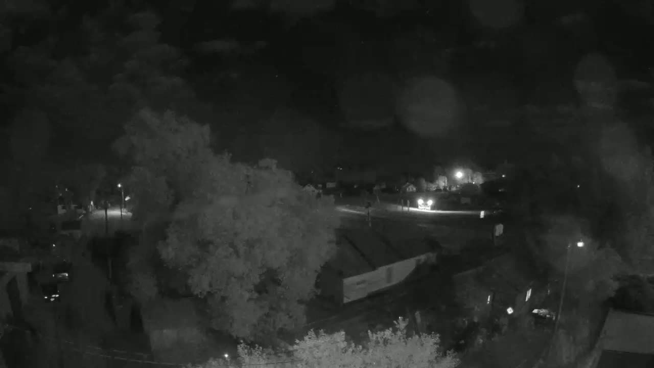 A nighttime, low-light view shows a sparsely lit street with some trees and buildings, possibly residential, under a dark, possibly cloudy sky.