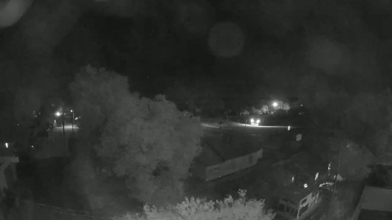 A nighttime, possibly rainy, view of a residential area with a vehicle's headlights visible in the distance, surrounded by trees and buildings.