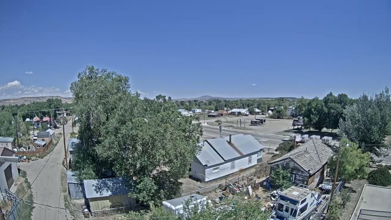 A high-angle view shows a small town on a sunny day with a few clouds, featuring residential houses, trees, and a road, with a distant mountain visible in the background.
