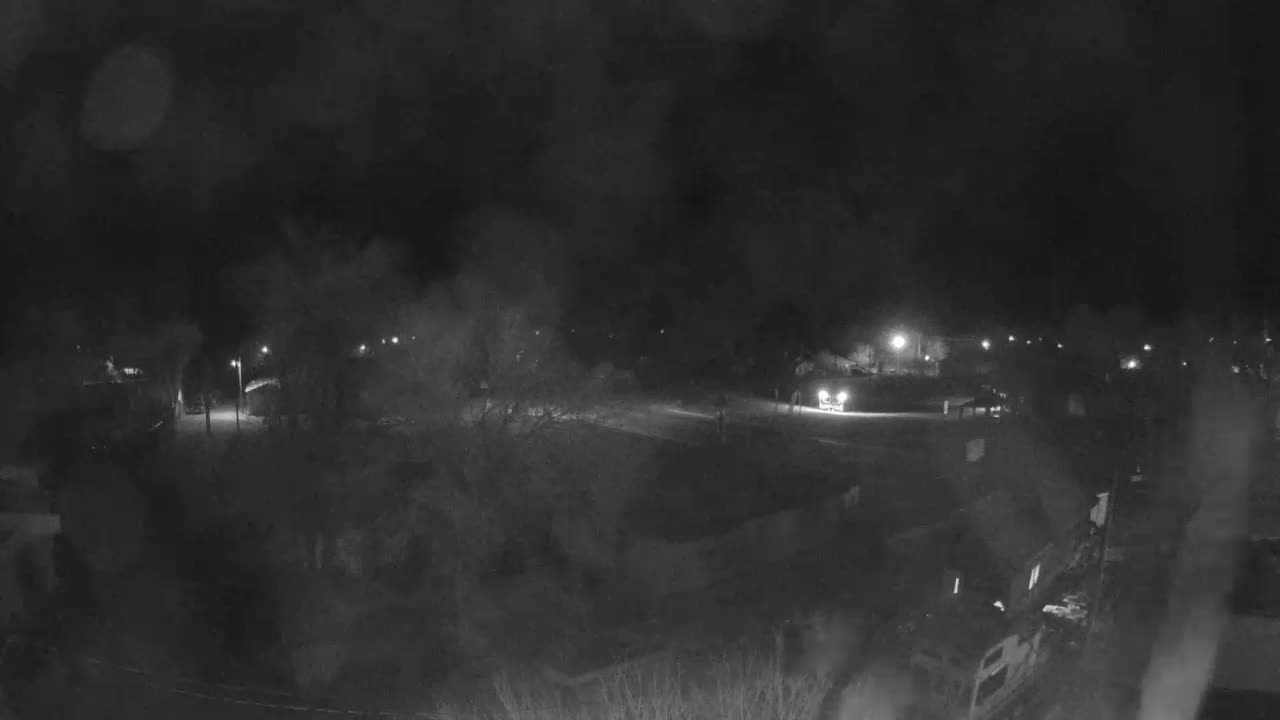 A blurry, low-light, grayscale outdoor scene at night, viewed through a rain-streaked lens, shows scattered streetlights and a vehicle with bright headlights on a distant road.