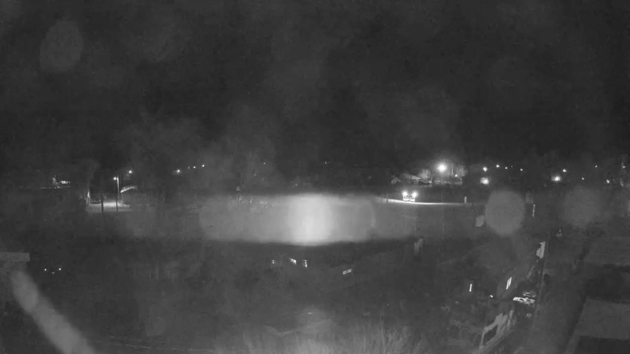 A blurry, grainy black and white image depicts a rainy night outdoors, with scattered streetlights and prominent vehicle headlights illuminating the dark scene and creating significant light flares due to moisture on the camera lens.