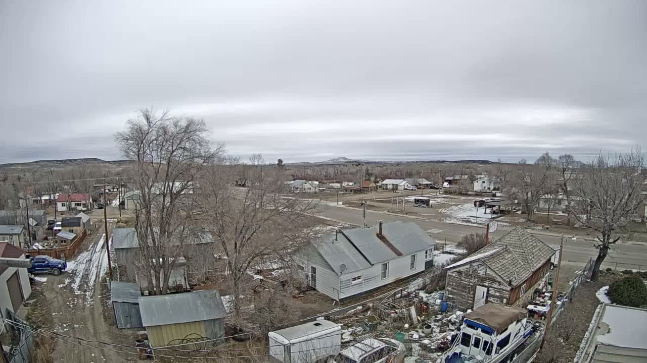An elevated view on an overcast day reveals a scattered rural or small-town landscape with various houses, bare trees, and patches of snow on the ground, under a gray sky with distant hills.