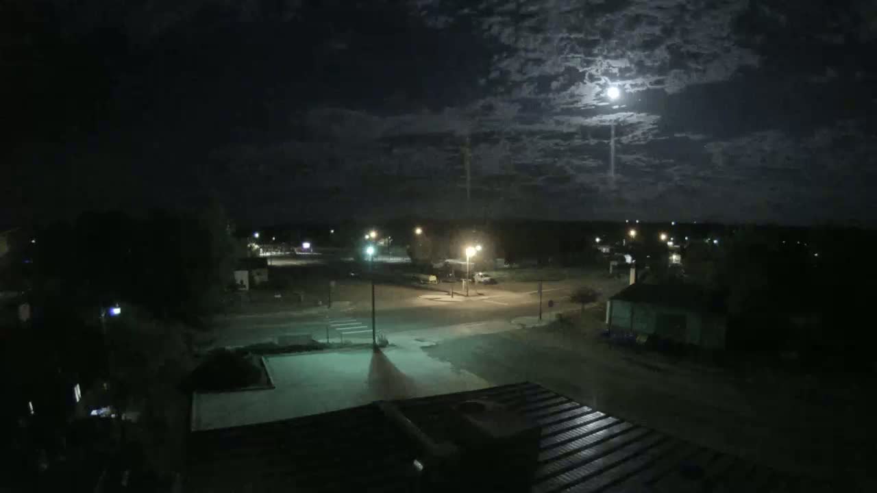 A nighttime view of a quiet street intersection in a small town under a partly cloudy moonlit sky.