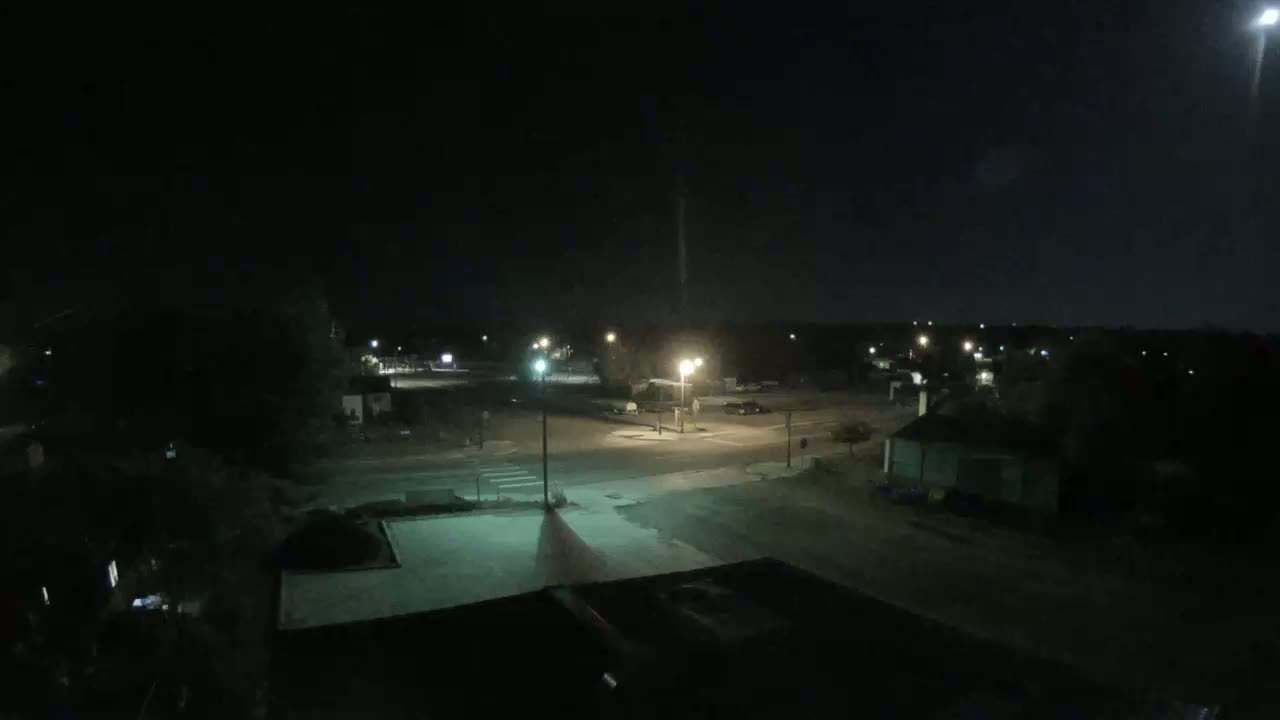 A nighttime view of a quiet street intersection in a small town under a partly cloudy moonlit sky.