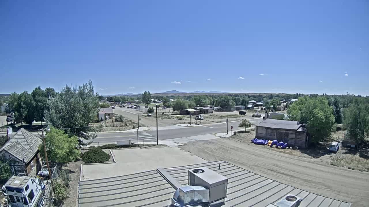 A high-angle, long shot on a clear, sunny day shows a small town with low buildings, roads, and some trees, with mountains visible in the background.