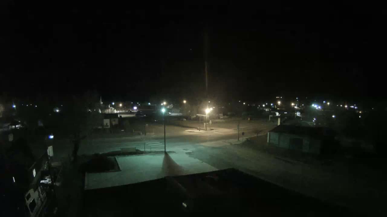 An overhead view reveals a dimly lit town at night under a clear, dark sky, with multiple streetlights casting beams onto roads and buildings.